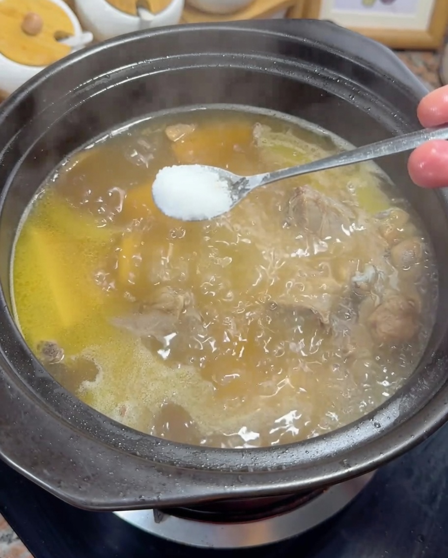 Añadiendo una cucharada de sal blanca a un caldo de pollo dorado hirviendo.