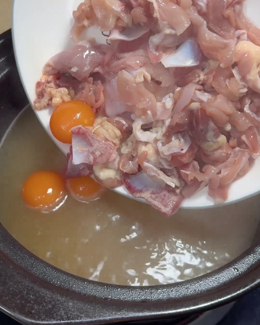 Carne de pollo cruda cortada fina y yemas amarillas siendo añadidas a una olla de caldo de pollo dorado hirviendo.