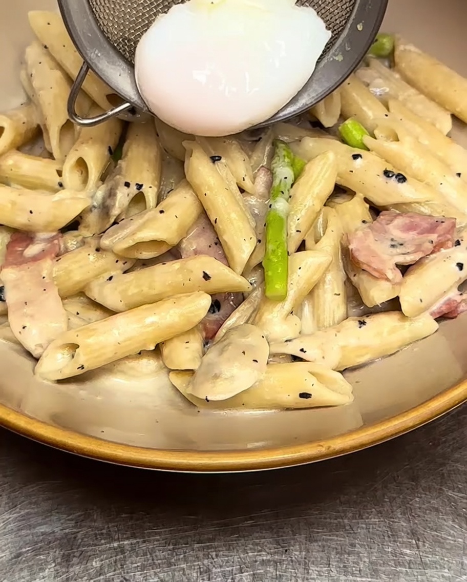 Pasta penne cremosa siendo servida en un tazón con un huevo pochado colocado cuidadosamente encima.