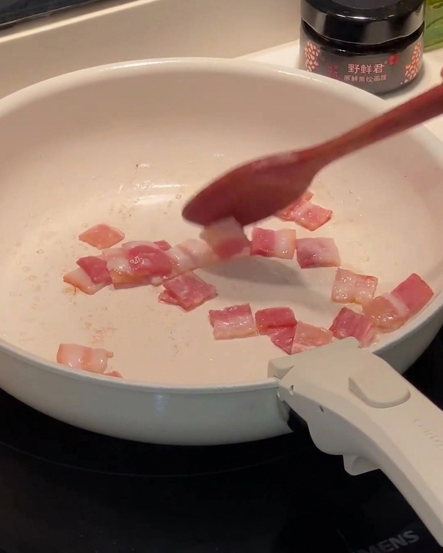 Tocino picado friéndose en una sartén blanca, con la grasa comenzando a acumularse en el fondo.