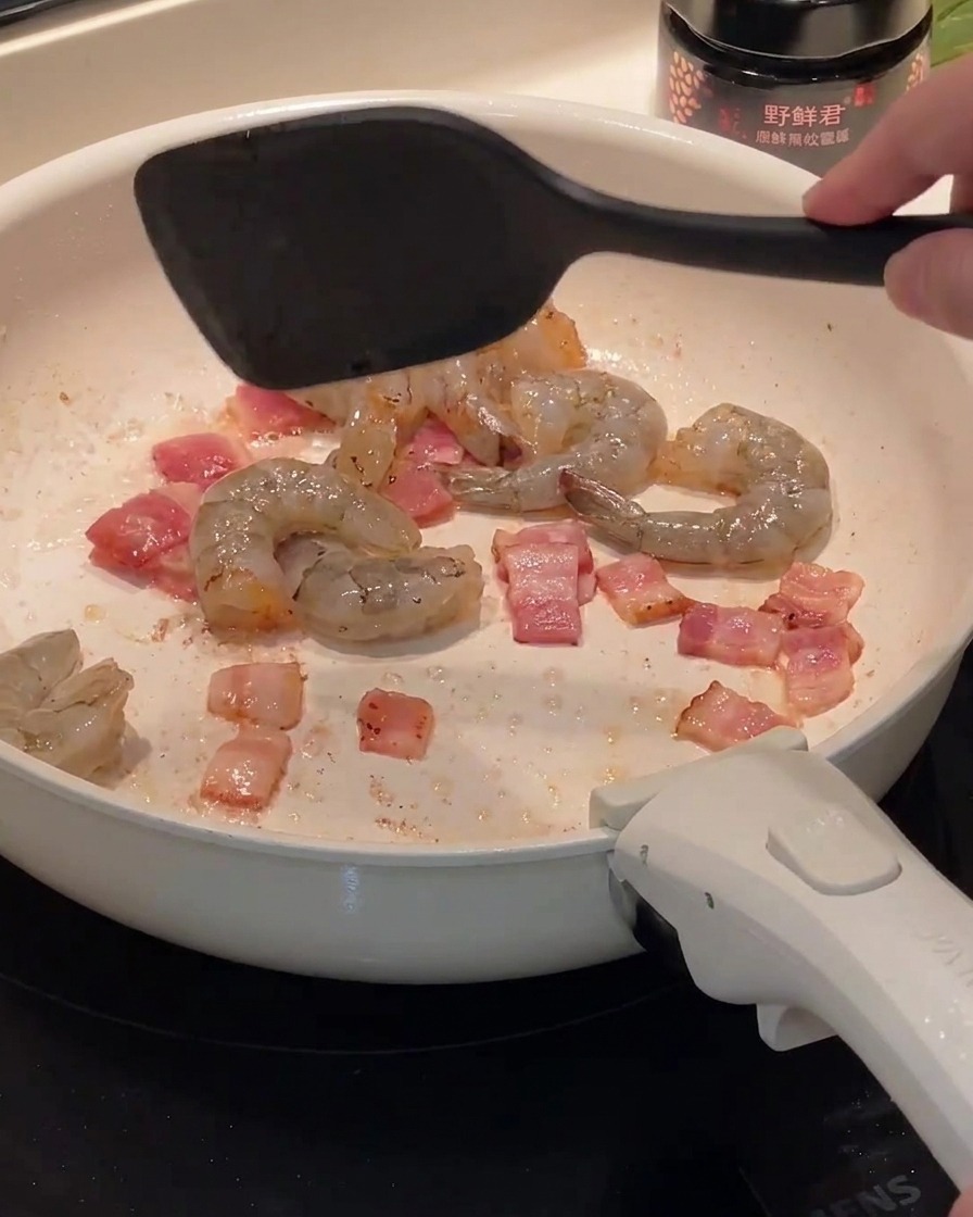 Camarones crudos siendo añadidos a una sartén que contiene trozos de tocino cocido.