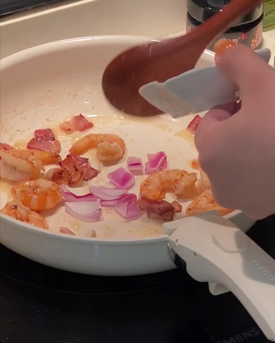 Cebolla roja picada siendo añadida a una sartén con tocino y camarones salteados.