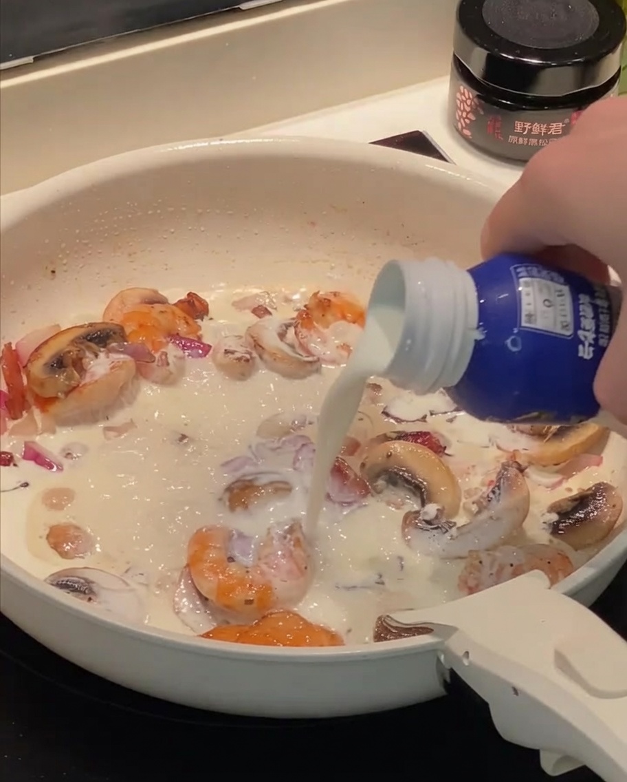 Crema de leche y leche siendo vertidas en la sartén con camarones, tocino y champiñones salteados.