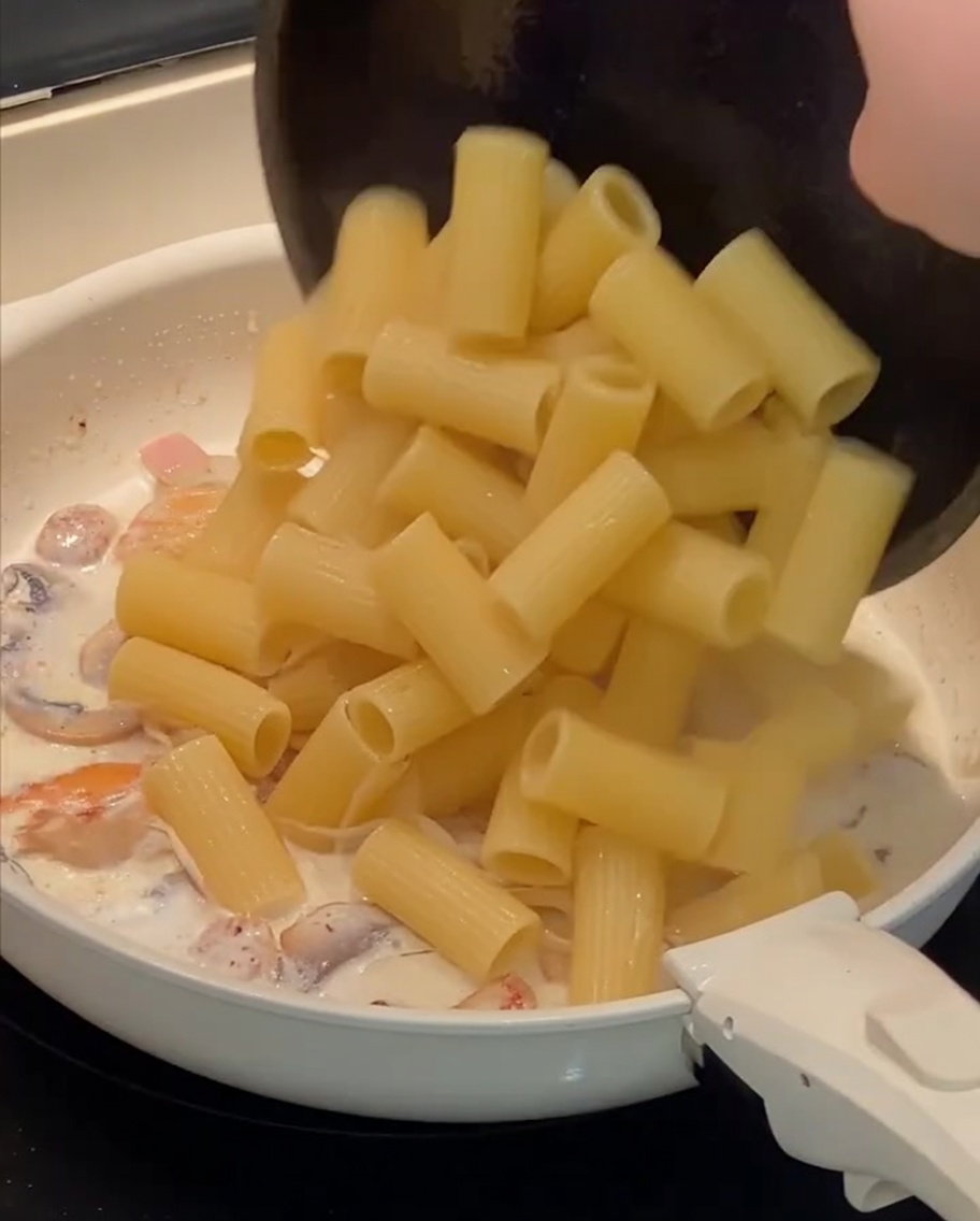 Pasta rigatoni cocida siendo añadida a la sartén que contiene la salsa cremosa y otros ingredientes.