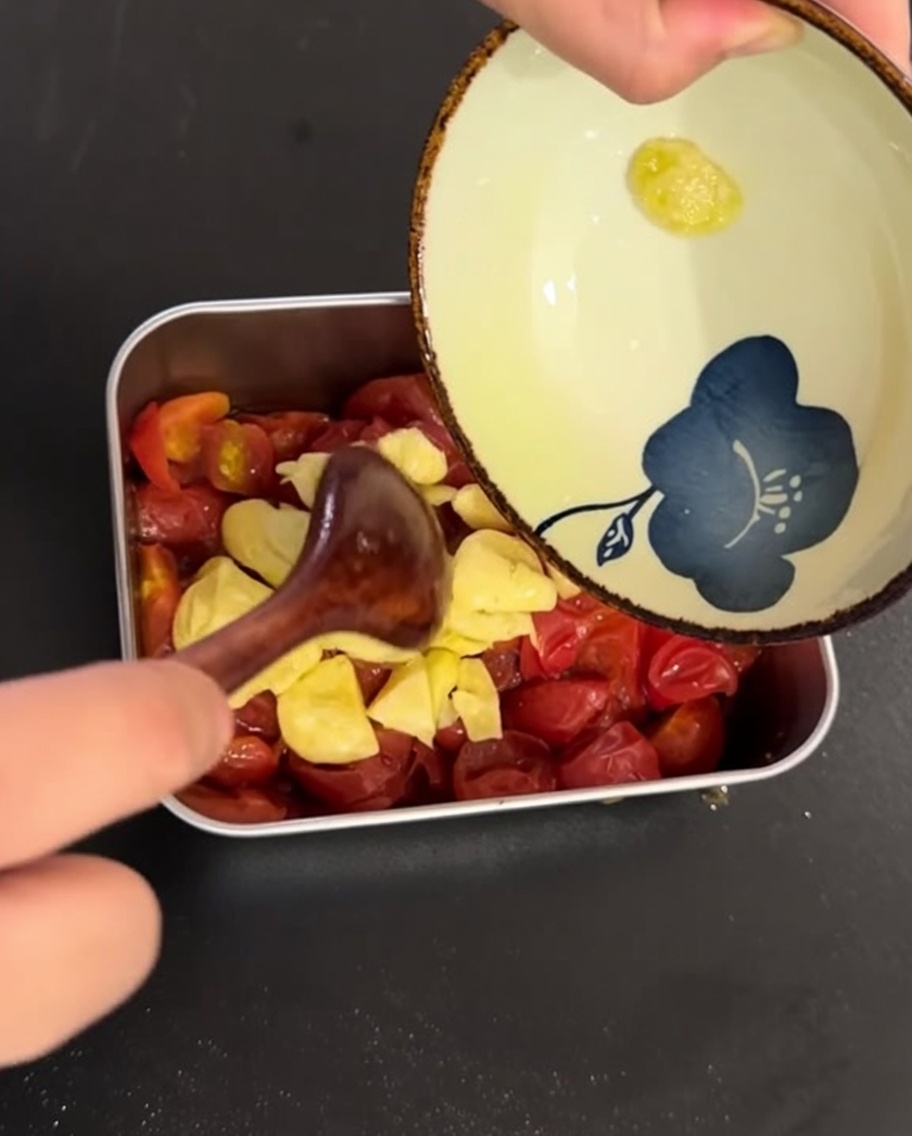El aceite infusionado con ajo y las láminas de ajo vertidas sobre los tomates cocidos en un recipiente metálico.