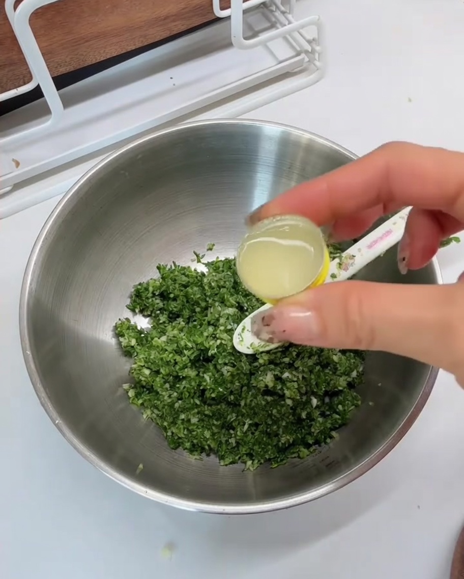 Jugo de limón vertiéndose desde una cuchara medidora en un tazón de metal que contiene la mezcla verde picada.