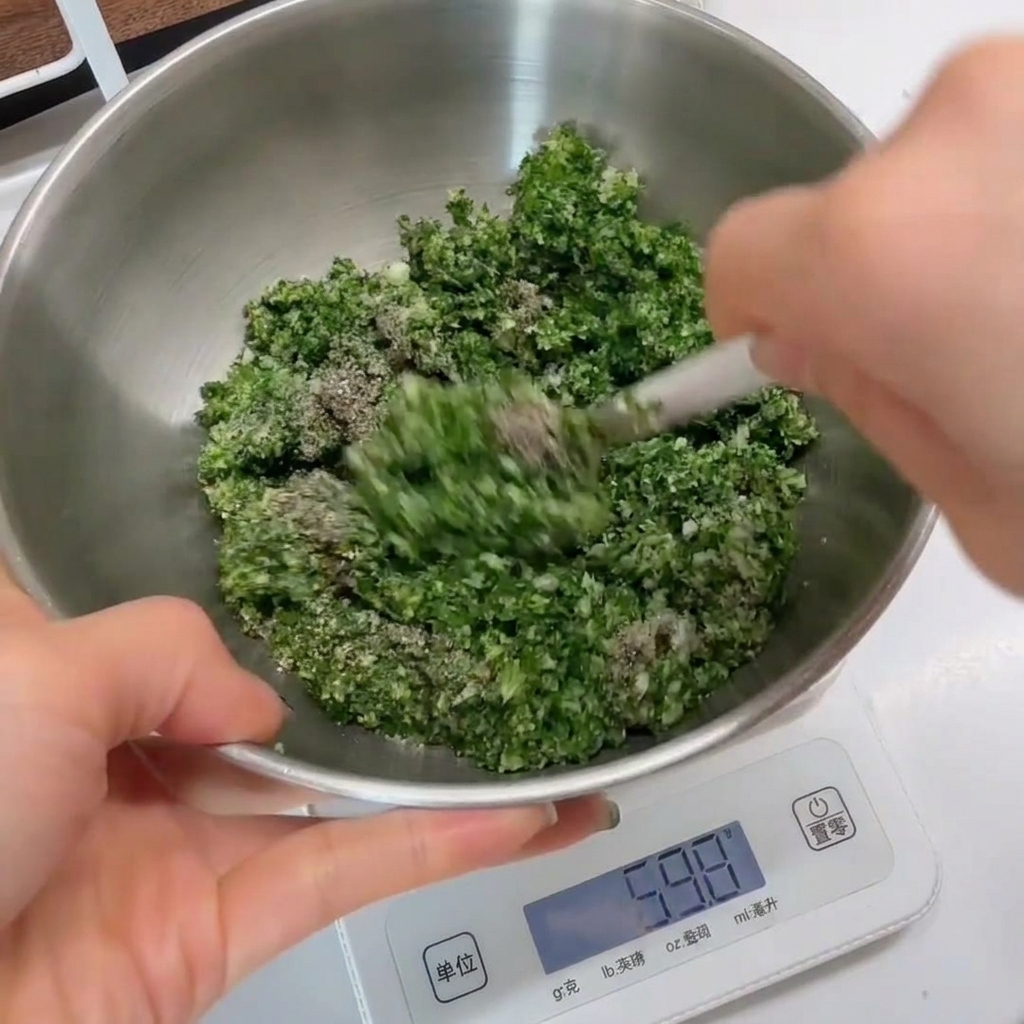 Primer plano de la mezcla de hierbas verdes siendo revuelta en un tazón de acero inoxidable con una espátula blanca.