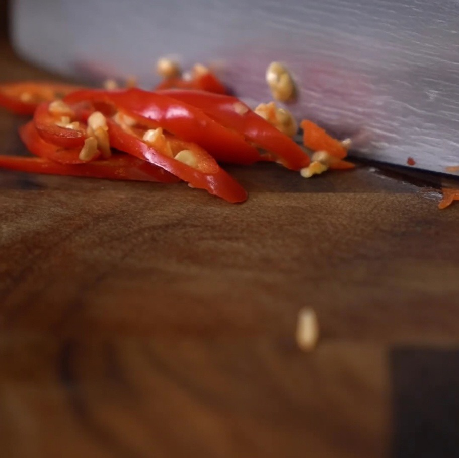 Un primer plano de un cuchillo rebanando chiles rojos brillantes en rodajas pequeñas sobre una tabla de madera oscura.