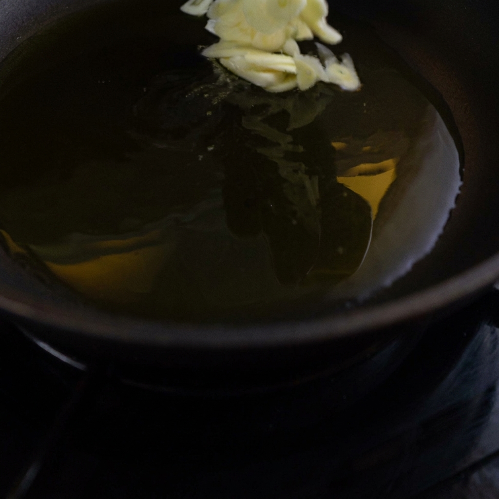 Láminas de ajo siendo añadidas a una sartén oscura llena de aceite de oliva caliente y brillante.