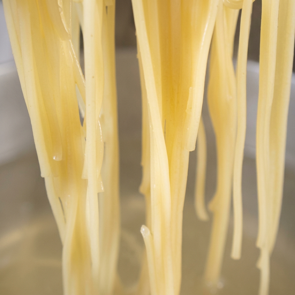 Fideos spaghetti cocidos siendo añadidos a una sartén con aceite de ajo dorado y chile rojo.