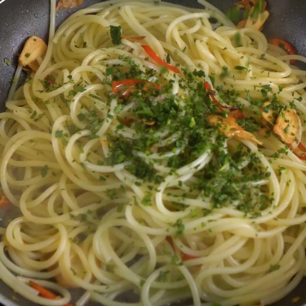 Perejil fresco picado siendo esparcido sobre el spaghetti y la mezcla de chile y ajo en la sartén.