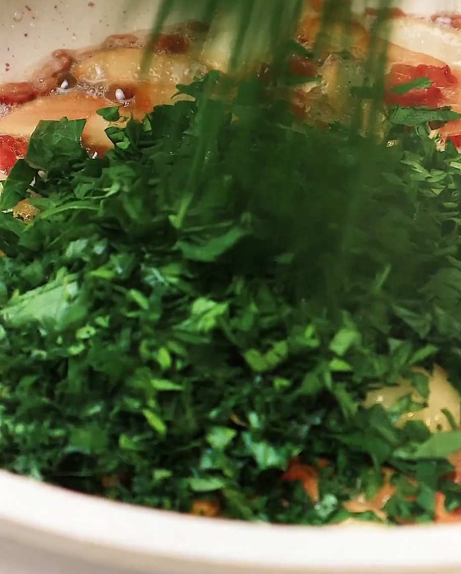 Perejil picado siendo añadido a la sartén con ajo y bacon, removiendo ligeramente para combinar.