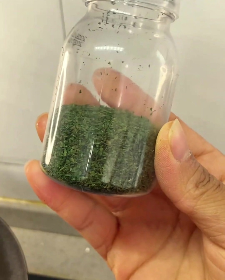 Una mano sosteniendo un pequeño frasco transparente parcialmente lleno con condimento de apio verde casero.