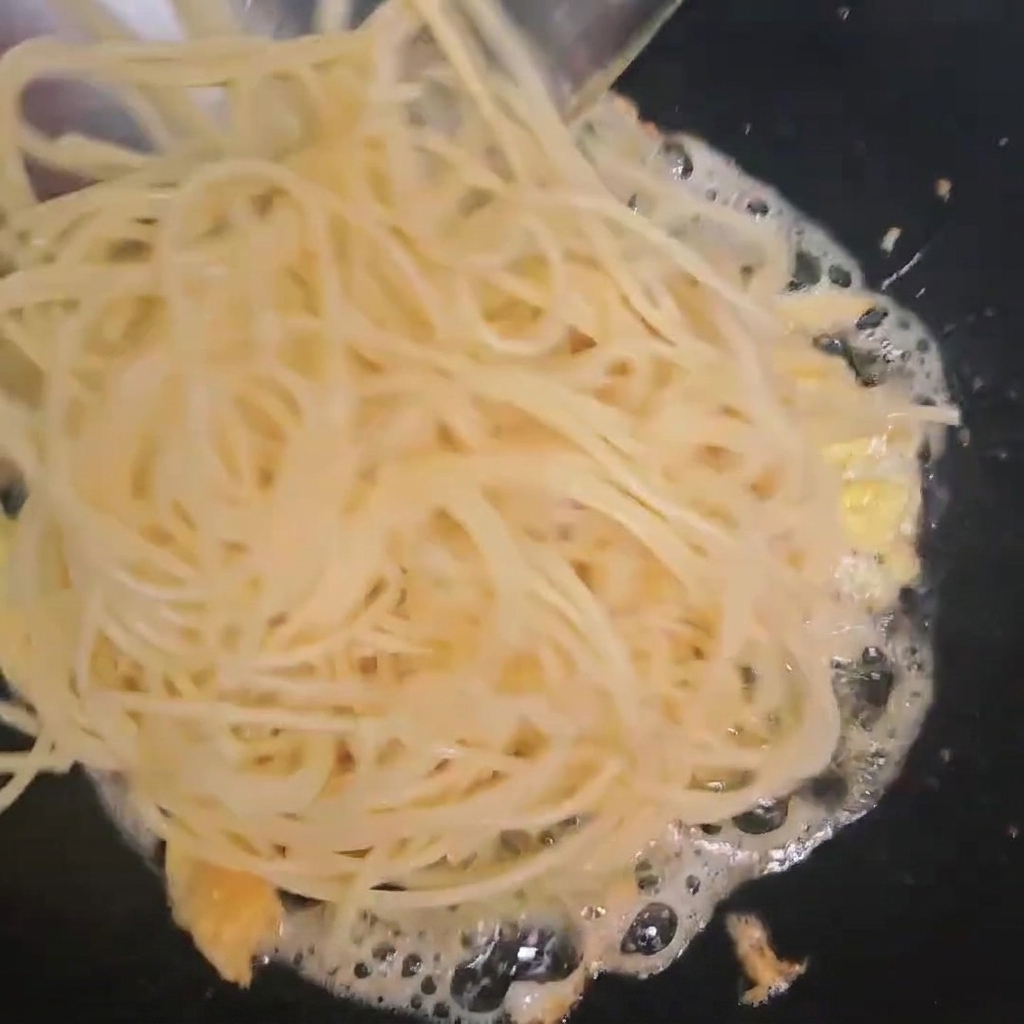 Espaguetis cocidos siendo añadidos a un wok que contiene huevos fritos.
