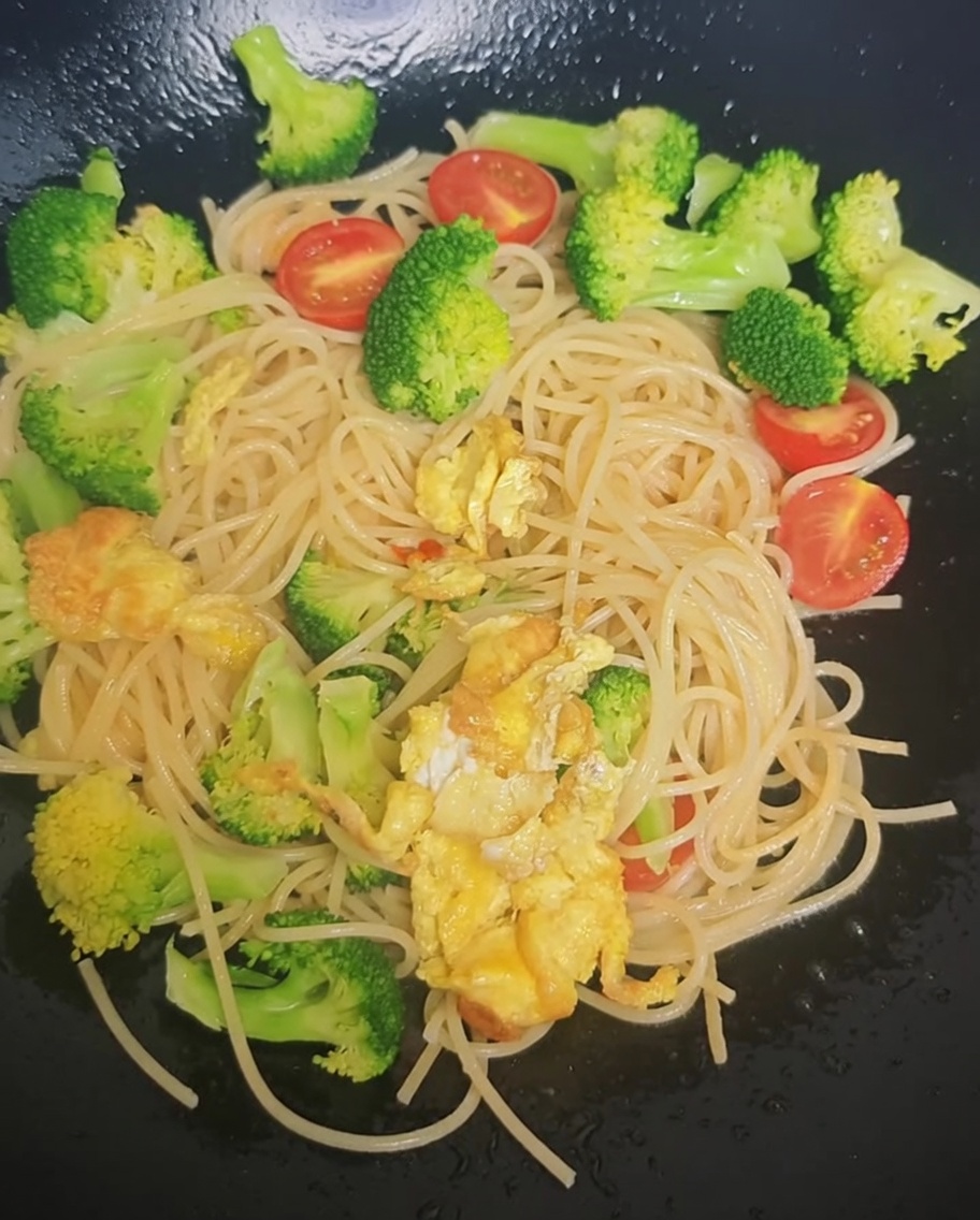Sazonador siendo espolvoreado desde un paquete sobre los espaguetis, brócoli y tomates cherry en un wok.