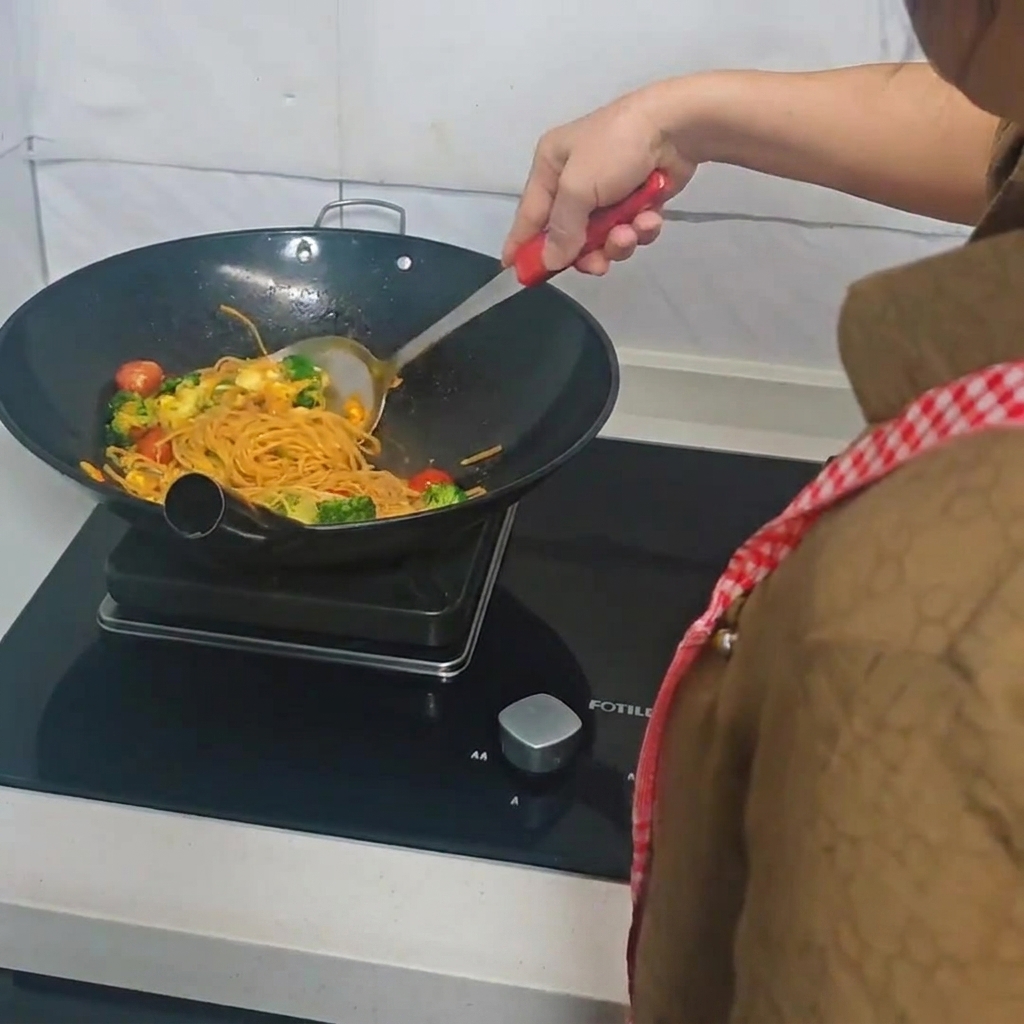 Una persona usando una espátula para saltear espaguetis, brócoli, tomates cherry y huevos en un wok.