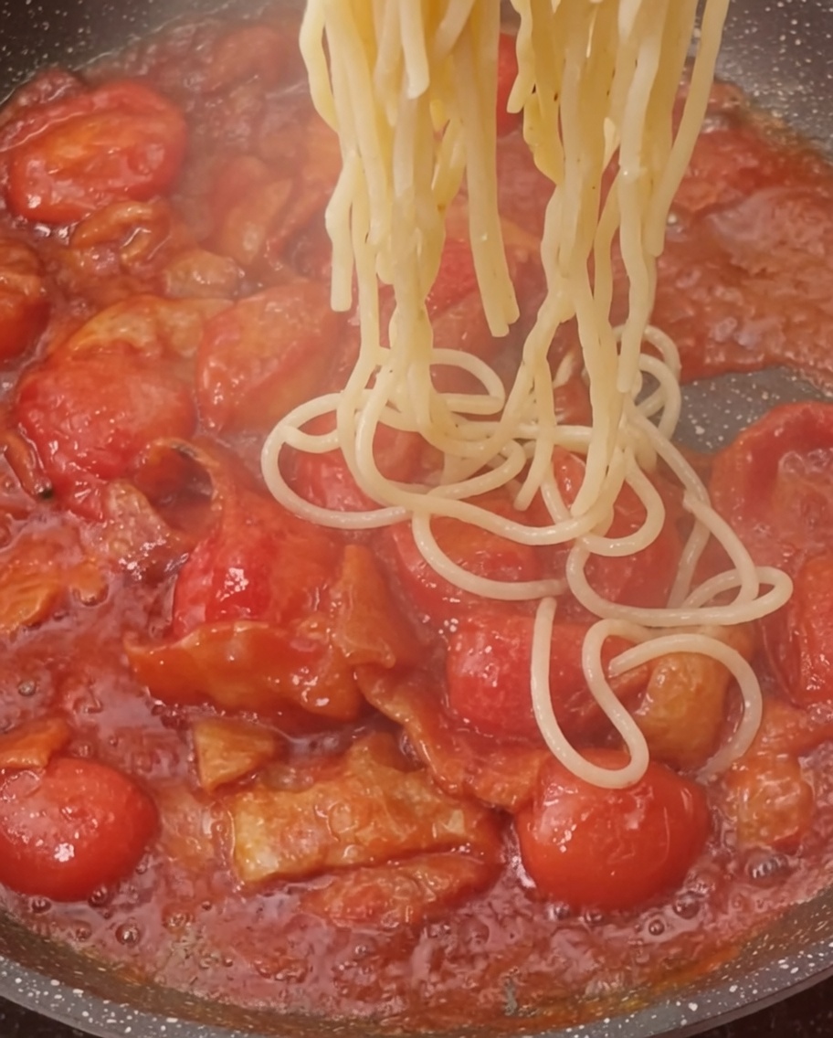 Pasta cocida siendo añadida a la sartén con la salsa de tomate y bacon.
