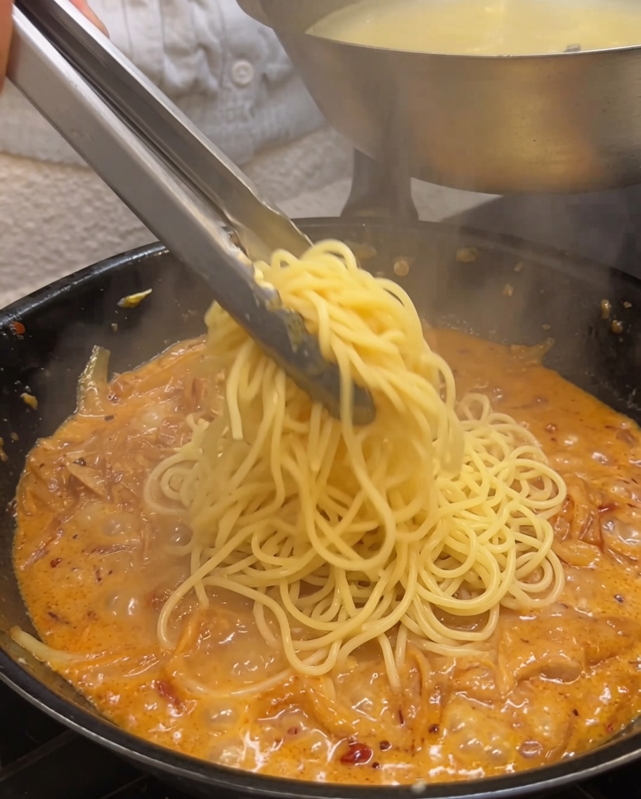 Espaguetis cocidos siendo mezclados en una salsa cremosa con pinzas.