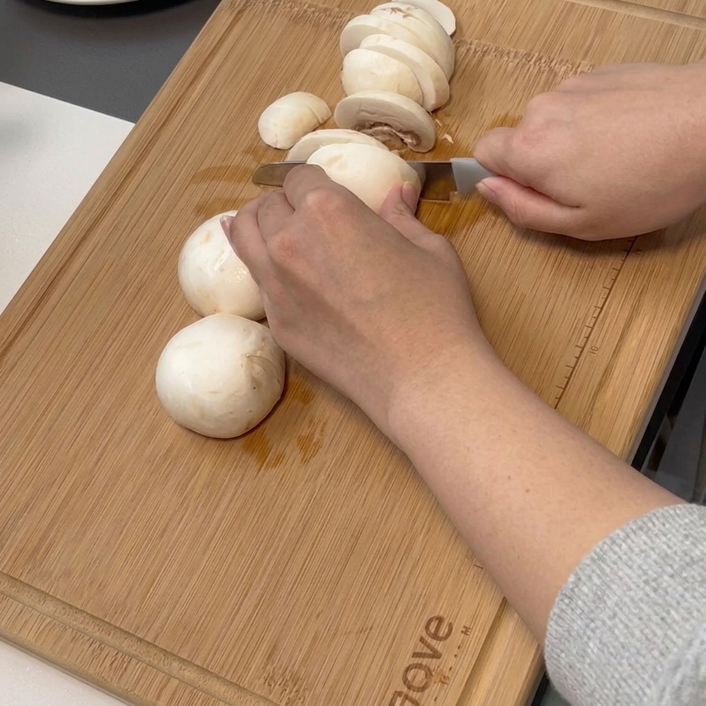 Una mano usando un cuchillo de chef para laminar champiñones sobre una tabla de bambú.
