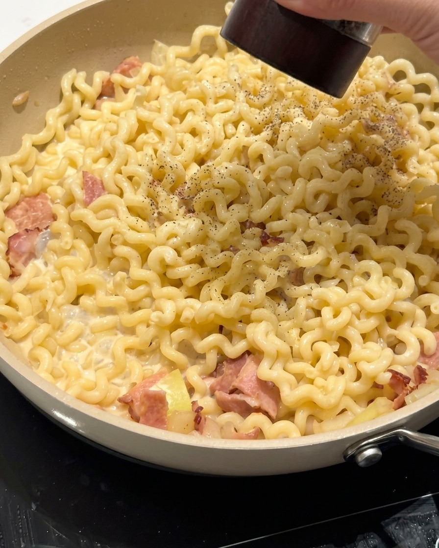 Pinzas mezclando fideos rizados cremosos en una sartén con bacon y salsa.
