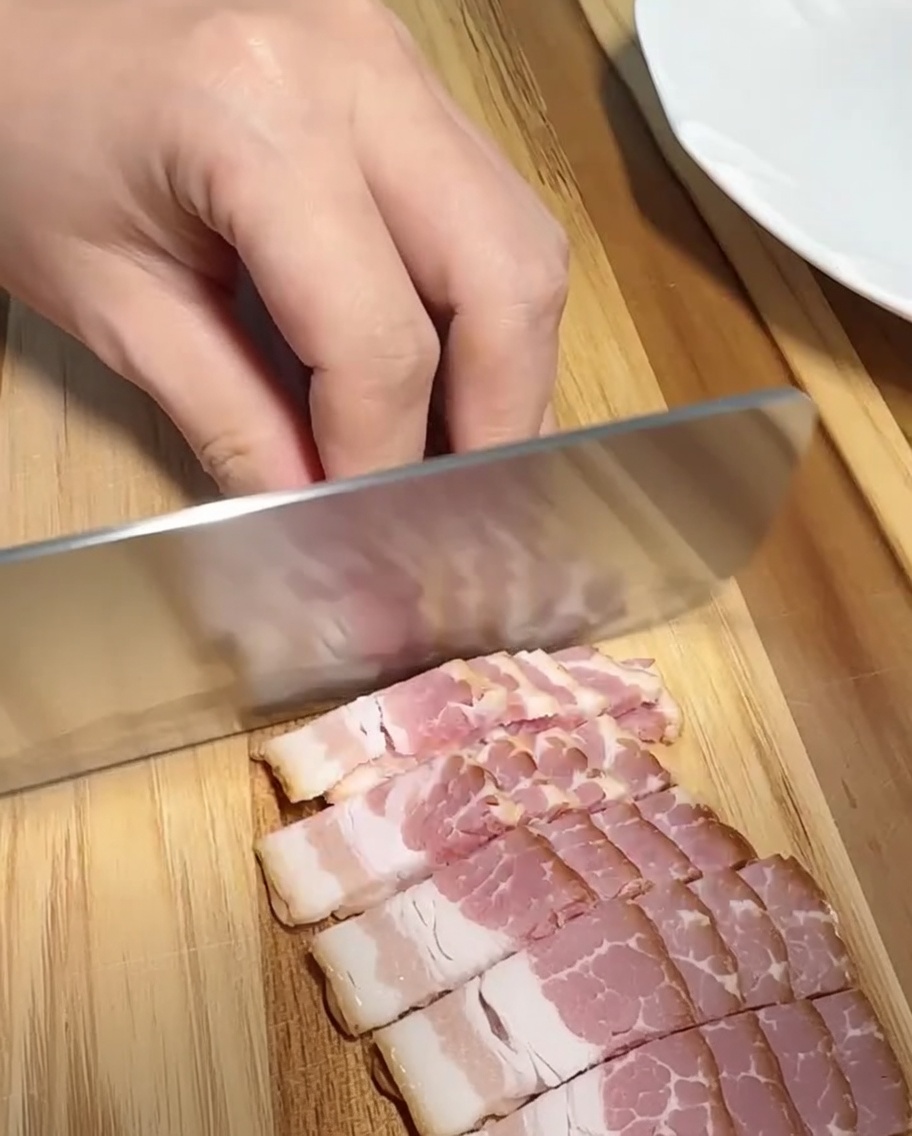 Primer plano de tiras de tocino siendo cortadas en trozos pequeños y uniformes sobre una superficie de madera.
