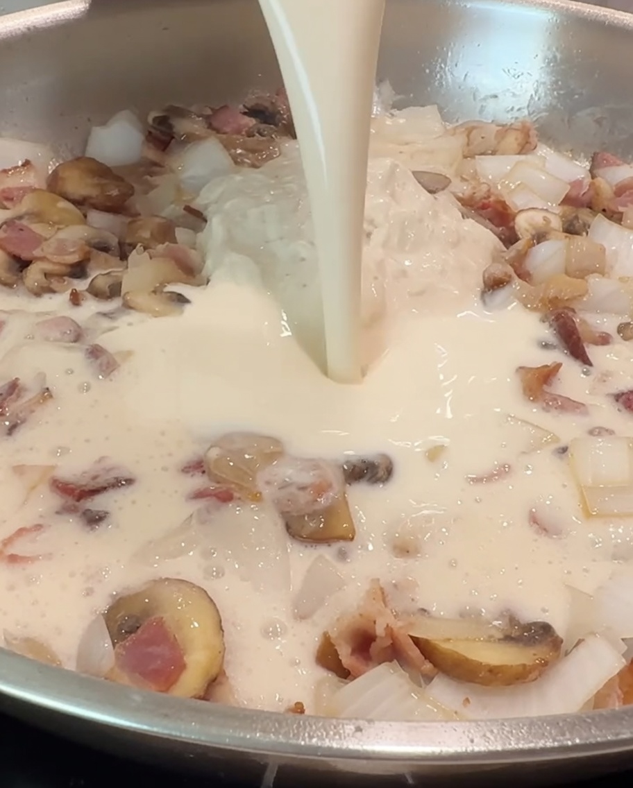 Una taza de leche siendo vertida en la sartén para mezclarse con la salsa Alfredo y los ingredientes salteados.