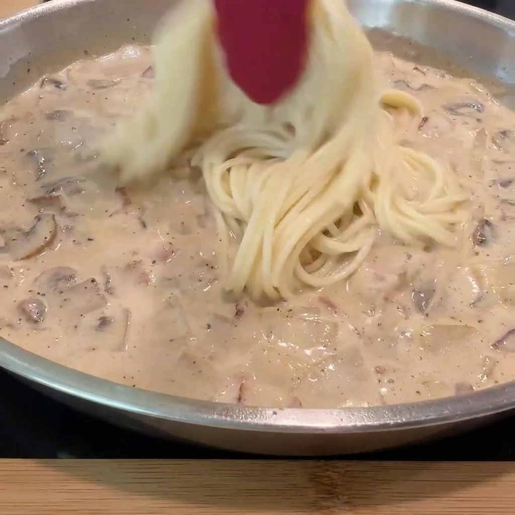 Fideos de espagueti cocidos siendo añadidos a la sartén con salsa cremosa e ingredientes.