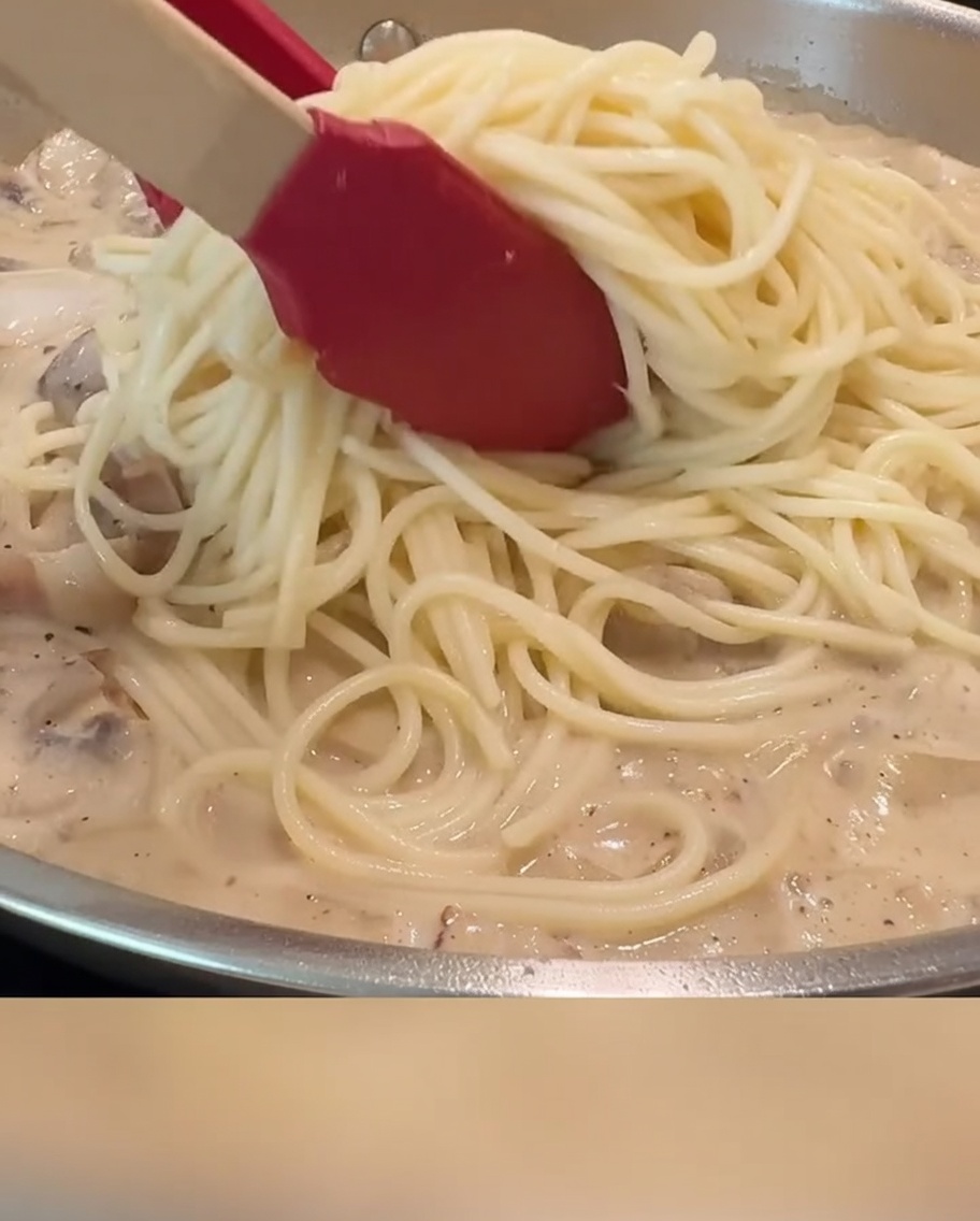 Espaguetis siendo mezclados con la salsa cremosa, el tocino y los champiñones en una sartén usando pinzas rojas.