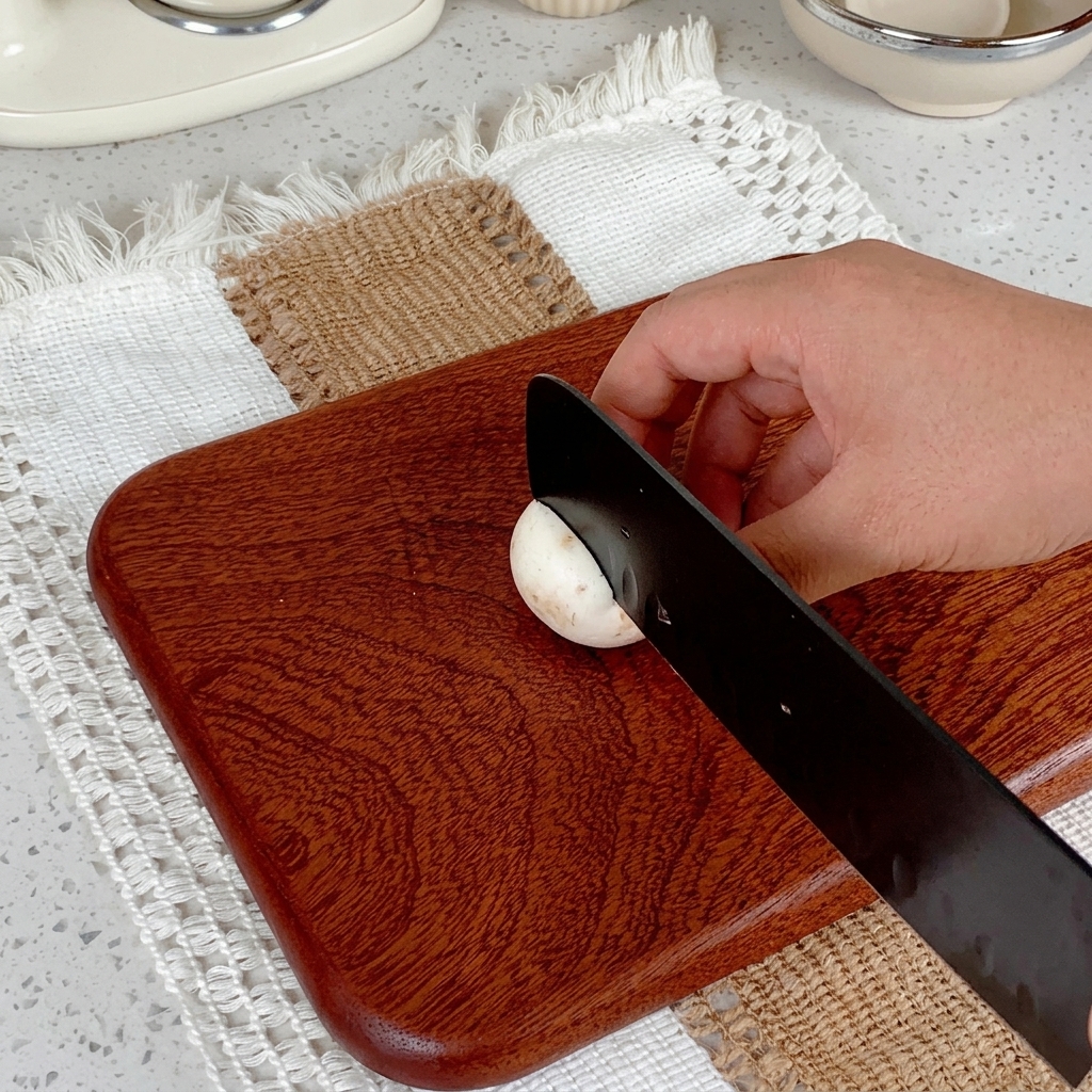 Una mano usando un cuchillo negro para rebanar un champiñón blanco sobre una tabla de madera.