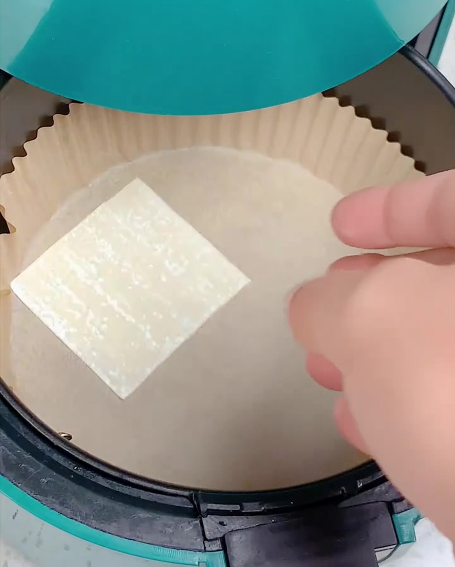 Una mano colocando una lámina de wonton cuadrada sobre papel pergamino en la freidora de aire.
