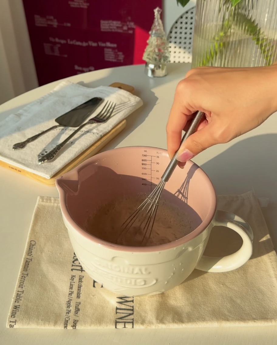 Primer plano de una mano batiendo levadura en agua tibia dentro de una jarra medidora rosa.