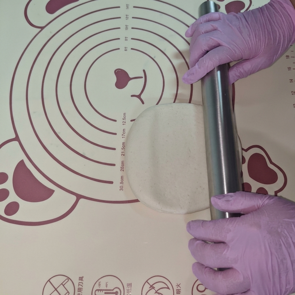 Una porción de masa siendo estirada en un círculo plano sobre un tapete de silicona usando un rodillo.