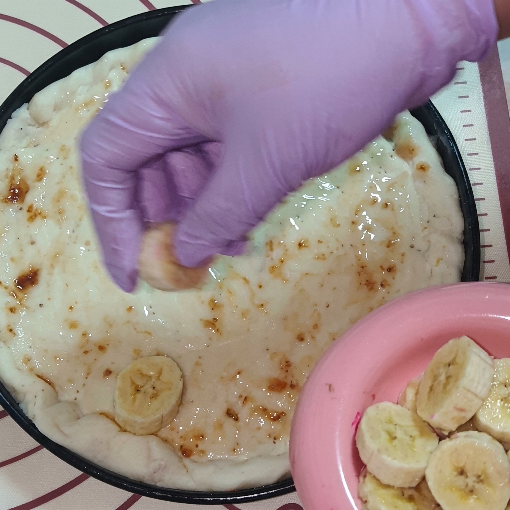 Una mano con guante morado colocando rodajas de fruta fresca, incluyendo plátano y pitahaya, sobre la masa cubierta de mermelada.