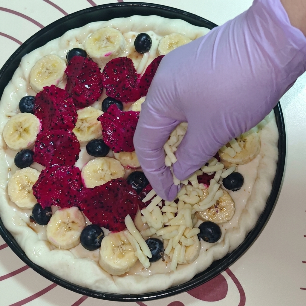 Vista superior de una mano con guante morado esparciendo queso mozzarella rallado sobre la pizza de frutas.