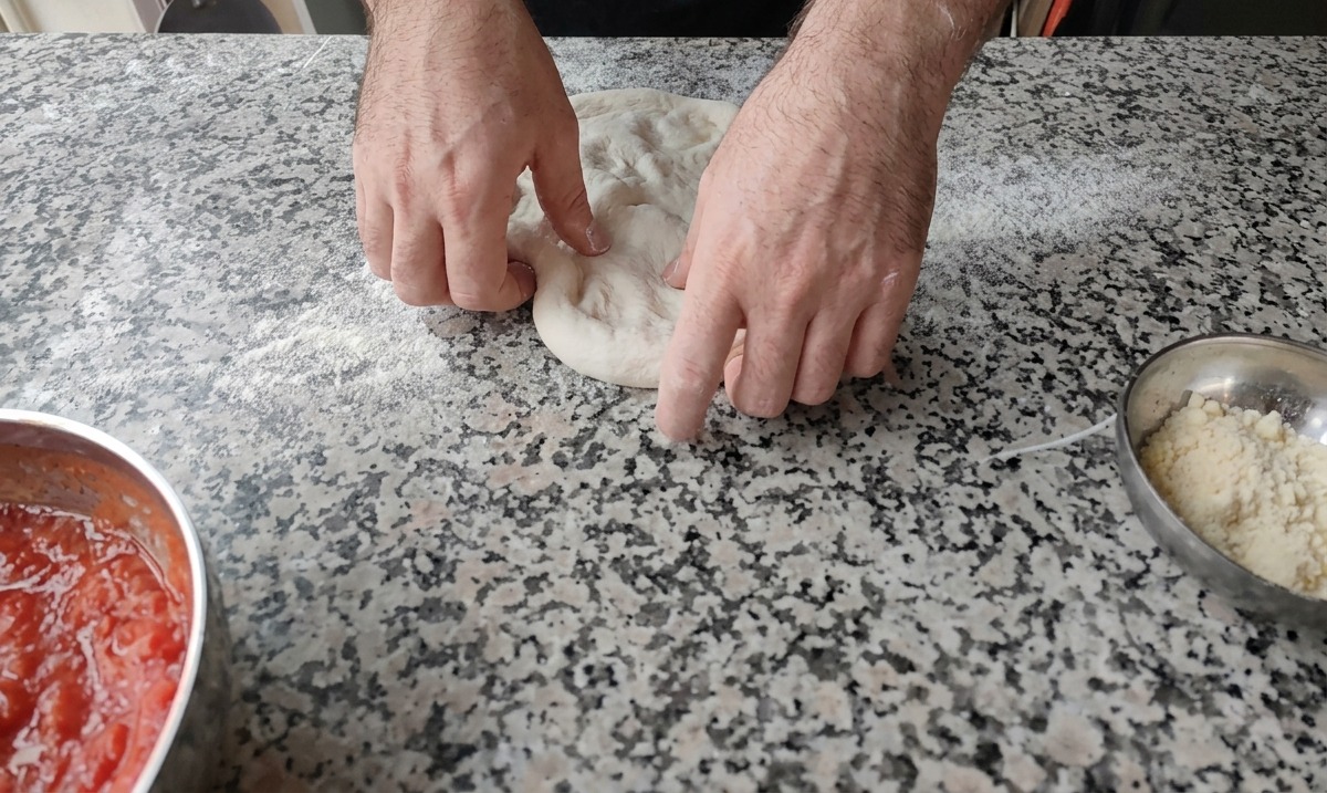 Dos manos presionando el centro de una bola de masa para pizza en una superficie de mármol enharinada.