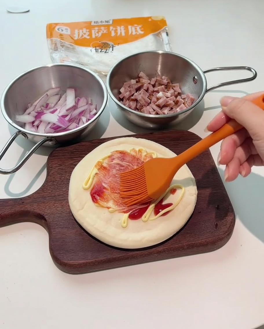 Un pincel de silicona naranja extendiendo salsas roja y blanca sobre la base de la pizza.
