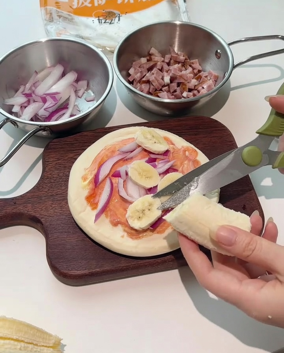 Tijeras de cocina cortando rodajas de plátano fresco directamente sobre la pizza.