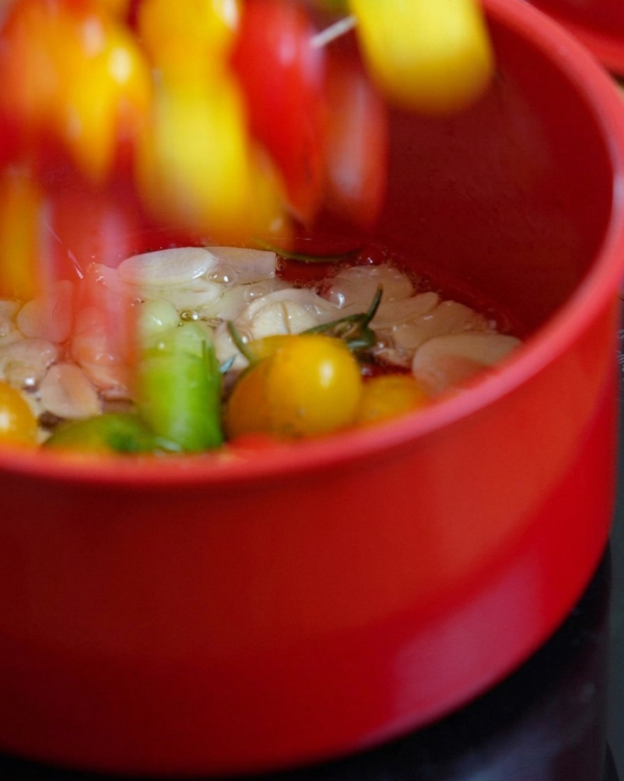 Tomates cortados añadiéndose a la olla con aceite caliente y hierbas.