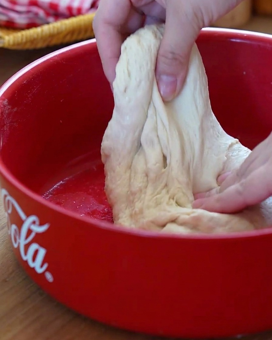 Manos realizando el estirado y pliegue en la masa dentro de un bol rojo.