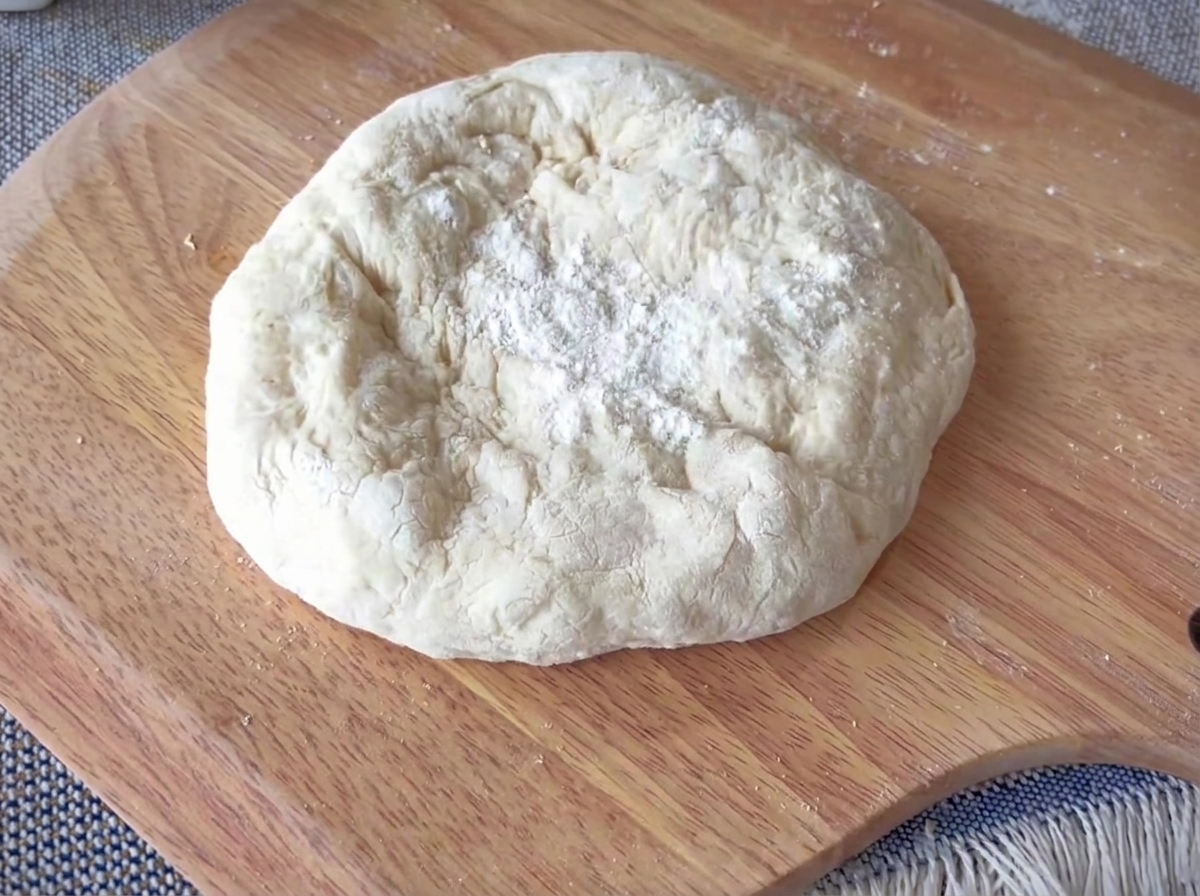 Una porción redonda de masa de pizza siendo estirada sobre una superficie de madera, con unos bordes gruesos bien definidos.