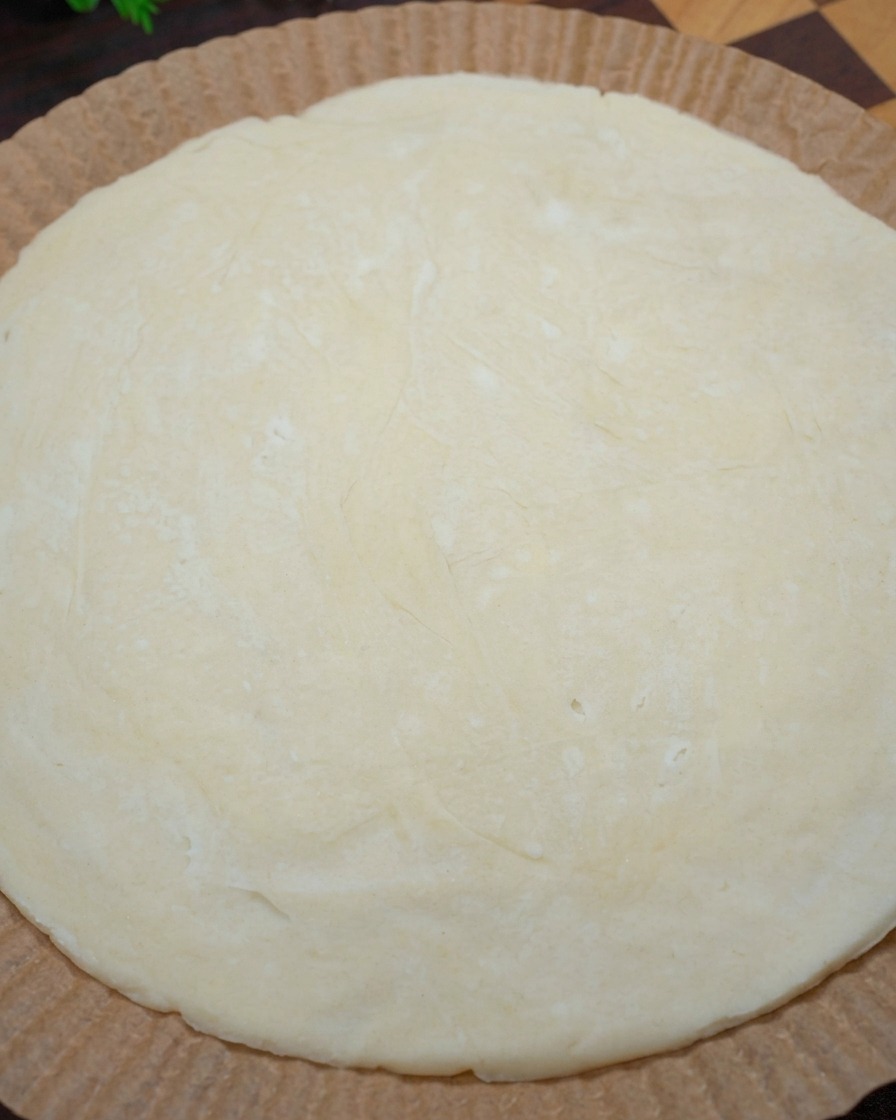 Primer plano de unas manos dando forma a los bordes de un panqueque de cebolleta crudo sobre papel vegetal para formar una base de pizza.