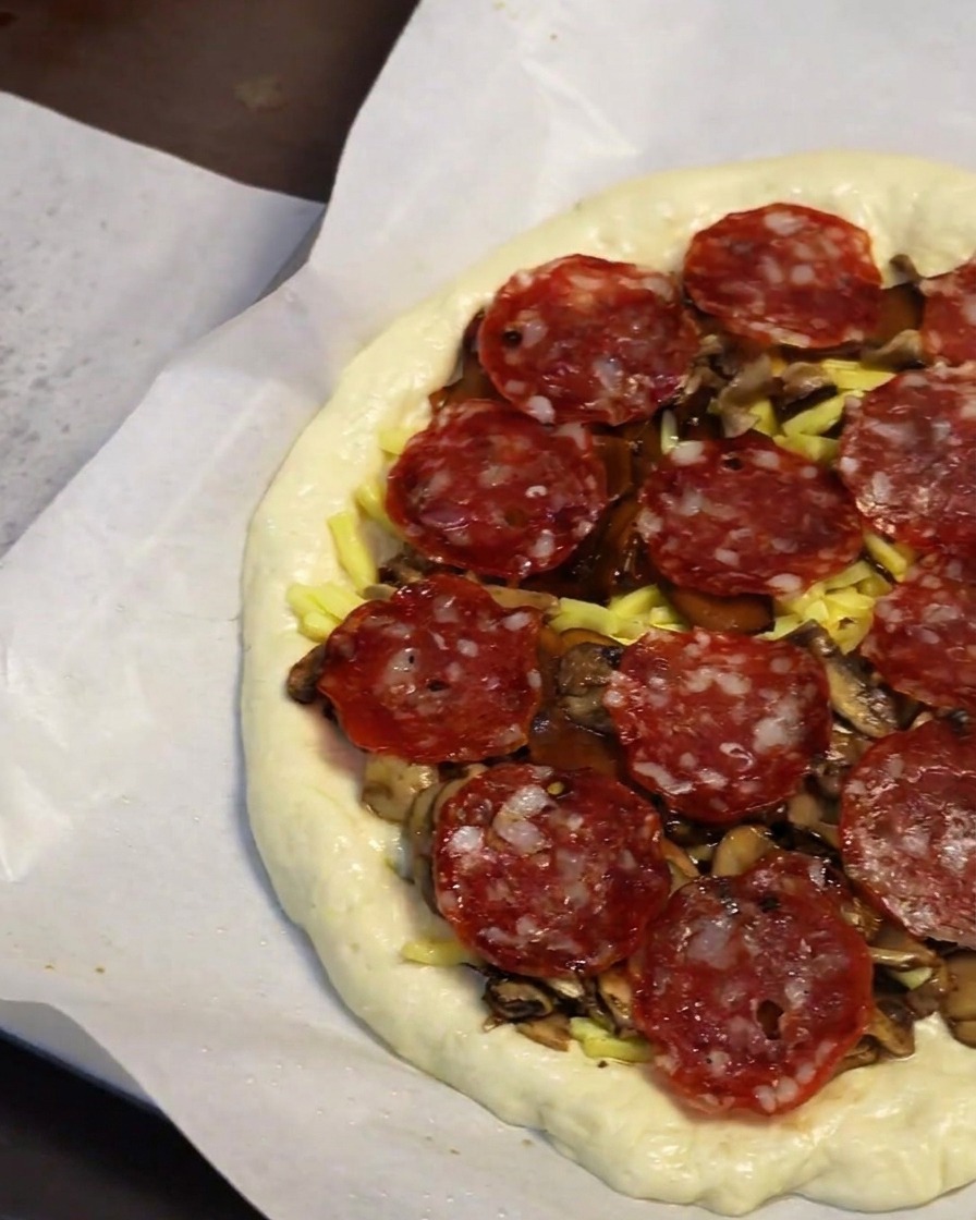 Primer plano de una mano colocando rodajas de pepperoni sobre los champiñones y queso en la masa.