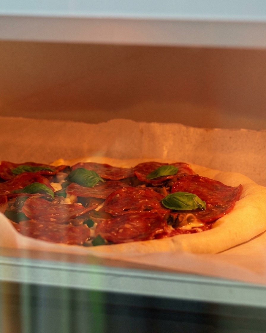 Una pizza cruda con queso, champiñones y pepperoni siendo colocada cuidadosamente en un horno caliente.