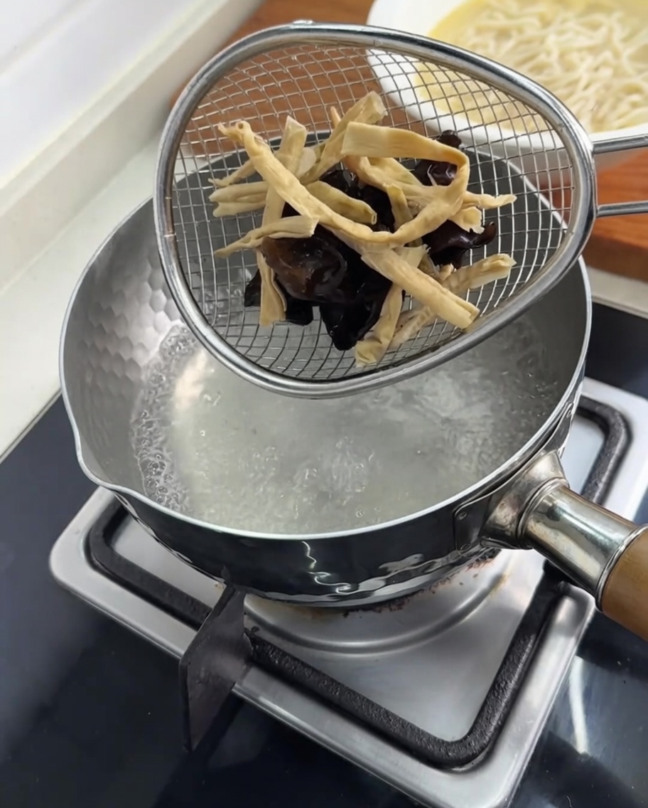 Hongos y brotes de bambú siendo blanqueados en un colador dentro de una olla.