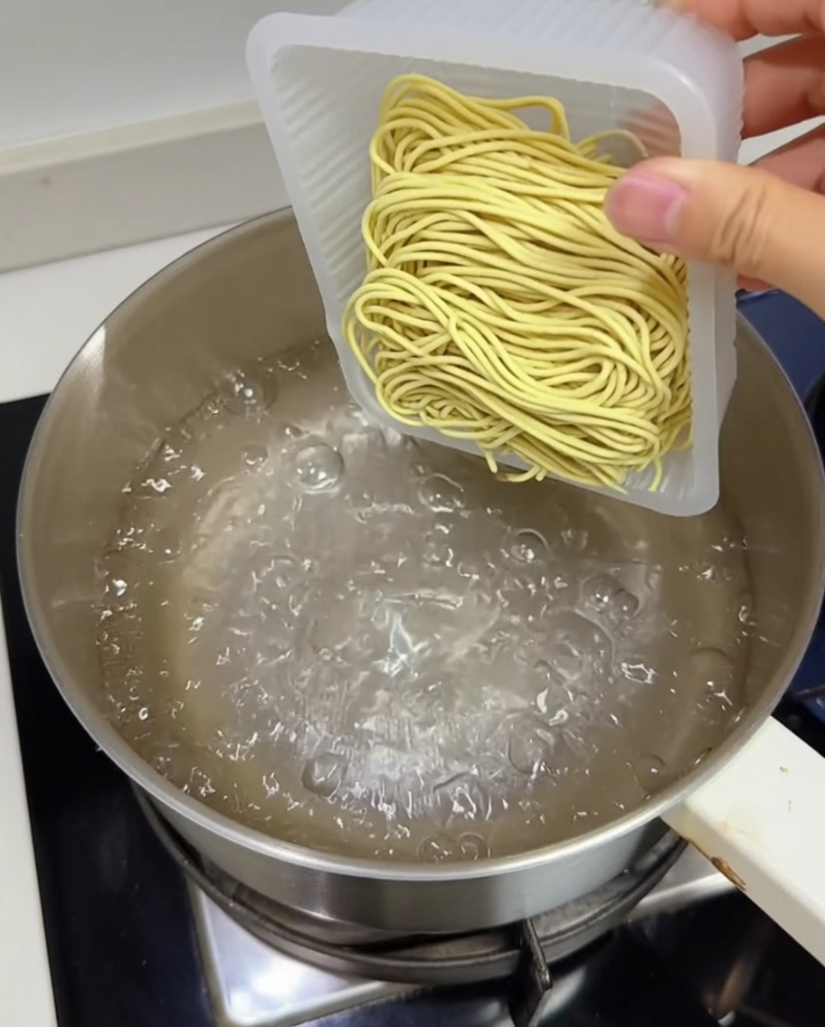 Un bloque de fideos de ramen amarillos frescos siendo añadidos a una olla con agua hirviendo.