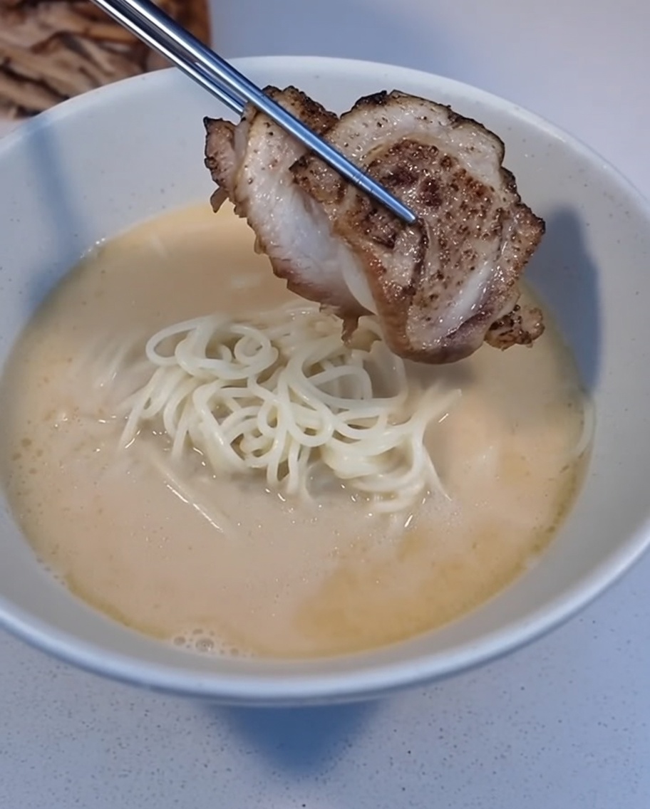 Una rebanada de Char Siu recién sellada colocándose sobre un tazón de ramen con pinzas.