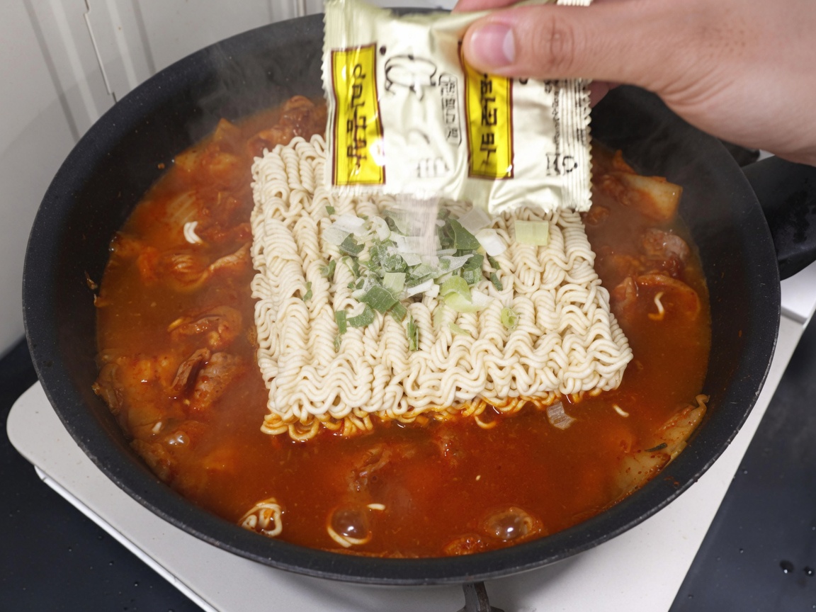 Paquete de condimento vertiéndose sobre los fideos ramen y el caldo en la sartén.