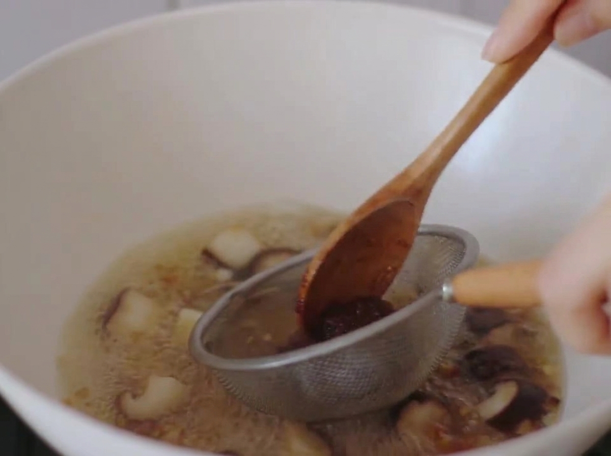 Pasta de miso siendo disuelta en el caldo hirviendo usando un pequeño colador de metal y una cuchara de madera.