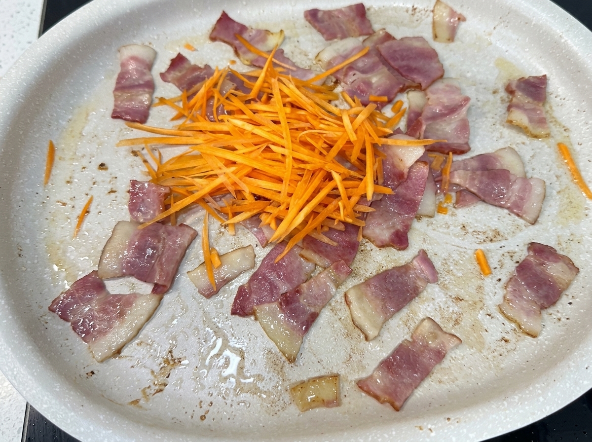 Zanahoria rallada, cebolla, repollo y brotes de soja añadiéndose a una sartén con trozos de tocino cocido.