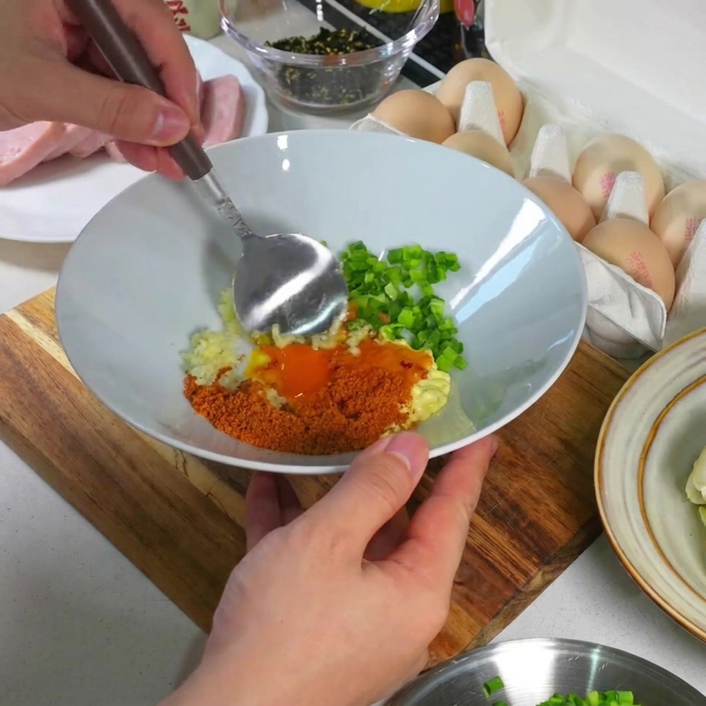 Primer plano de una cuchara mezclando la yema, el ajo, la cebolla y los condimentos en un tazón blanco hasta formar una pasta espesa y suave.